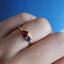 A two stone cluster ring with one blue round stone and a square princess cut deep pink stone on a thin gold band. 